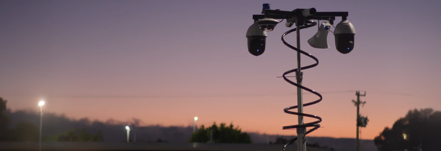 Commercial security cameras mounted on a pole at dusk, watching an outdoor lot under a purple and orange evening sky.