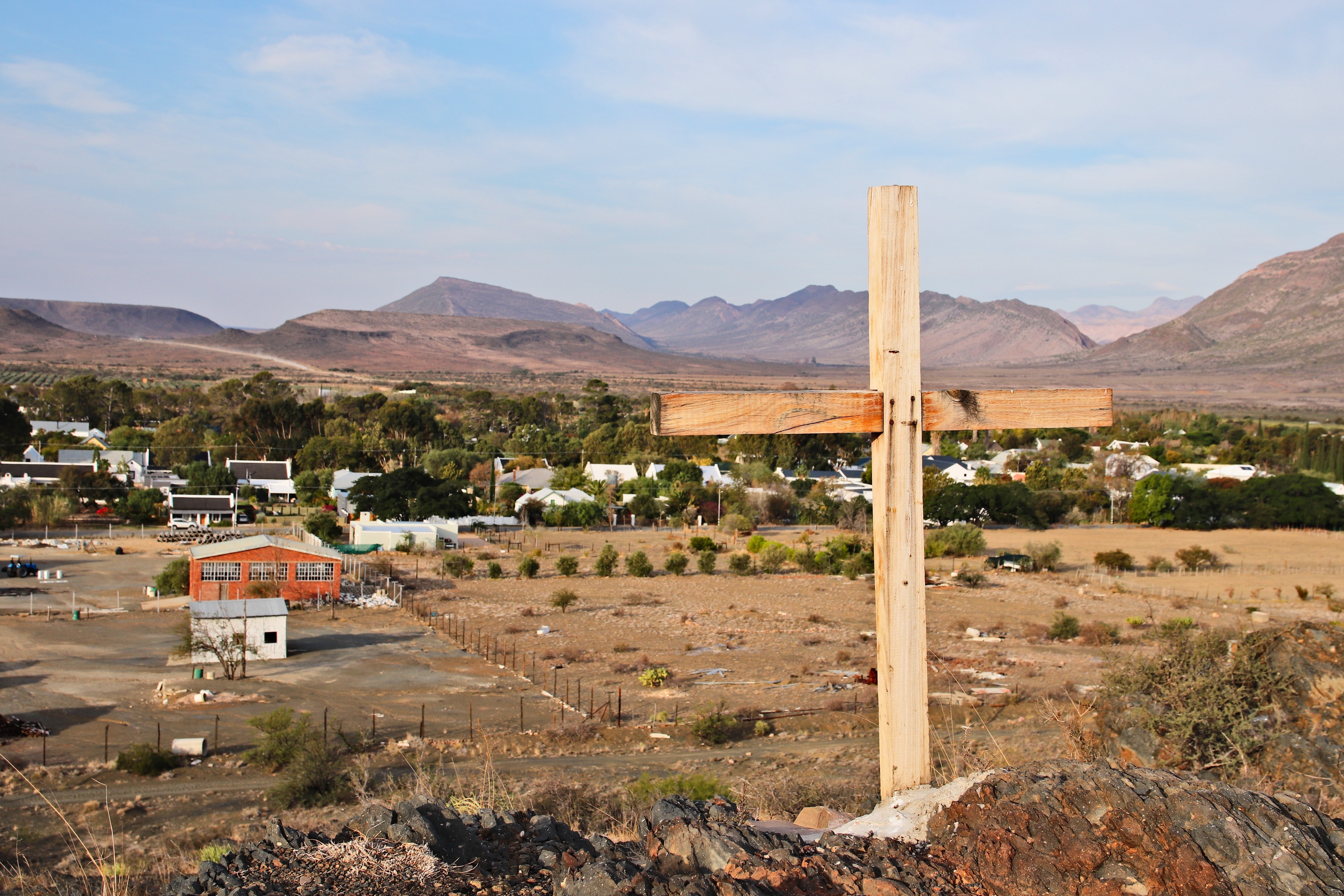 Wooden cross on a hill overlooking a small Southwest-style town with mountains in the distance.